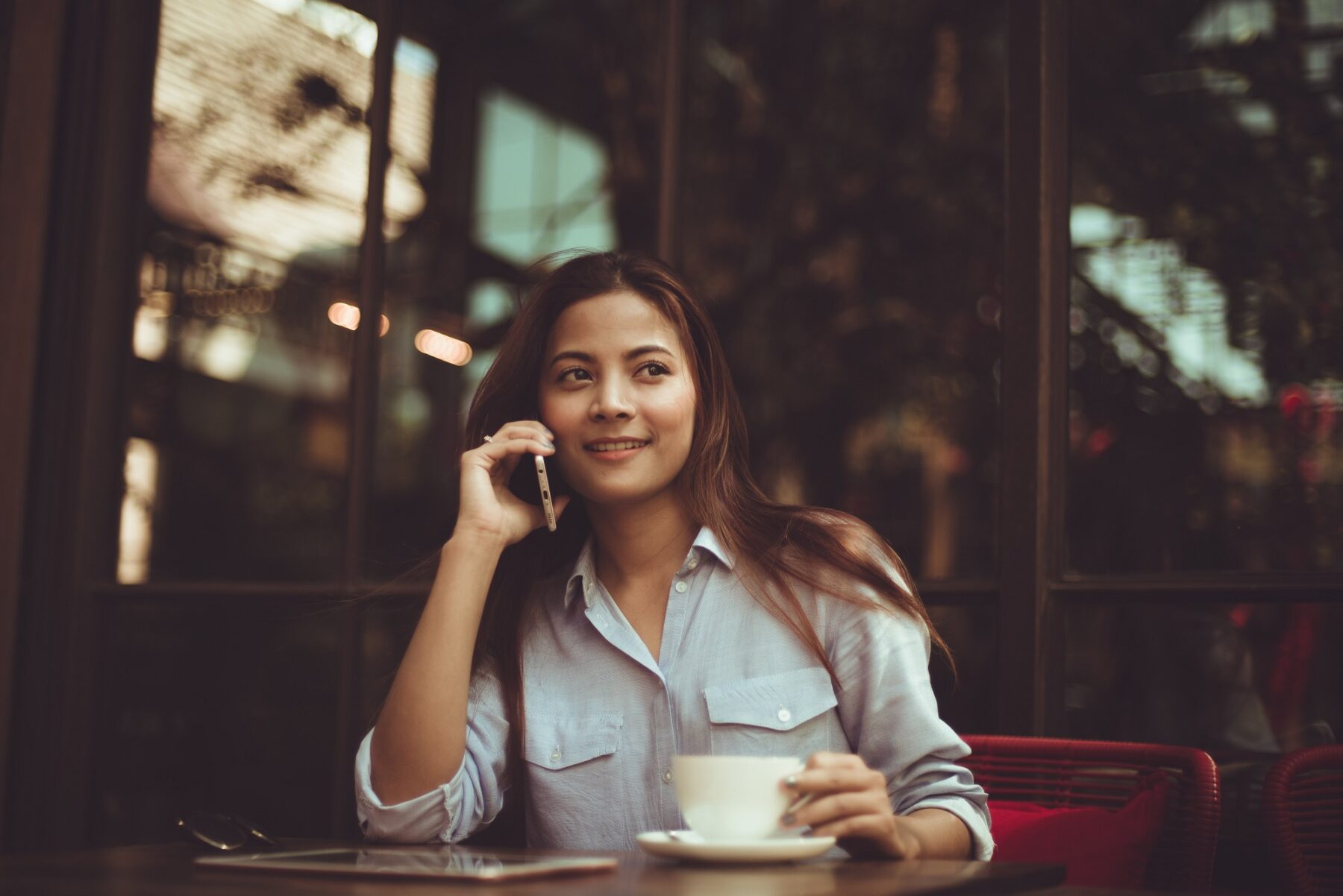 A woman sits at an outdoor café table, smiling while talking on her phone. She holds a white cup and saucer in one hand, with a tablet and plate in front of her. The background shows windows and reflected trees.